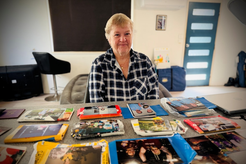 Debbie smiles as she sits in front of a benchtop of old photos, she has short blonde hair, blue front door, tv behind.