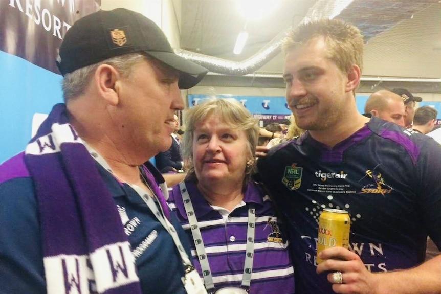 Smiling Steven, Debbie, and Cameron in purple and white football uniform embrace holding a beer, crowds behind him.