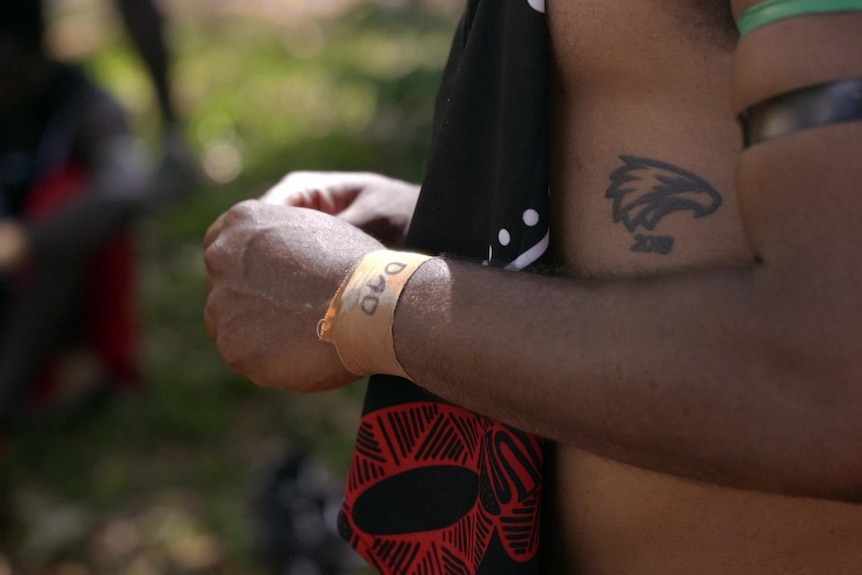 A man, shirtless, with an eagle tattoo on his ribcage and an orange wristband with the word 'Dad'.