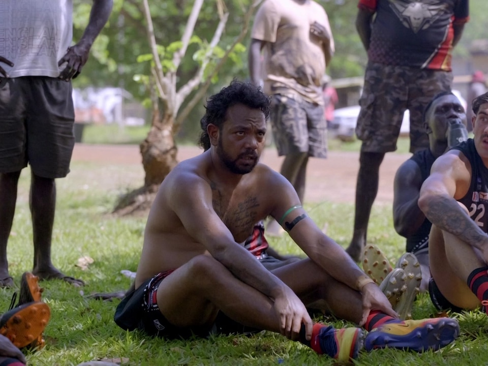 A man sits on the grass shirtless next to other footy players. Some other men are standing in the background.