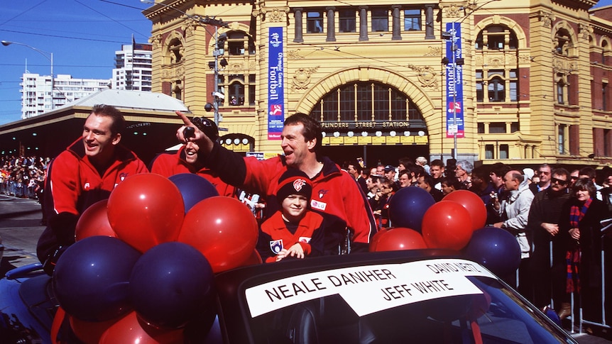 Two Melbourne AFL players and the coach and a child travel in a car, waving at the crowd at the AFL grand final parade.