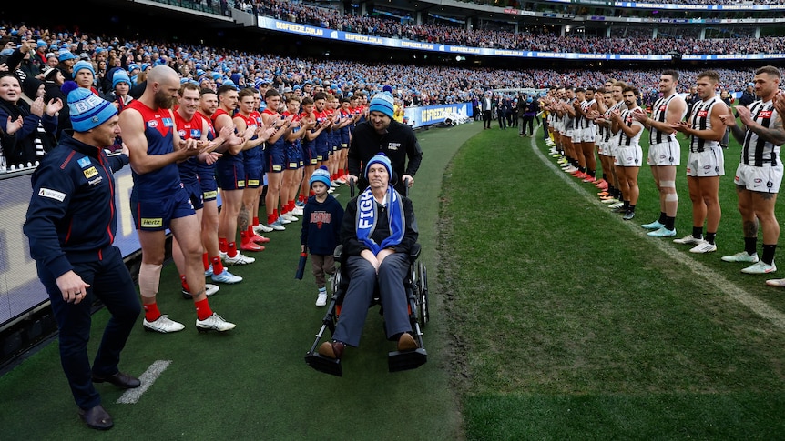 A former AFL player and coach is pushed on the grass at the MCG in a wheelchair, with Demons and Magpies players on either side.