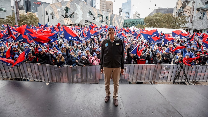 Former AFL player and coach Neale Daniher stands smiling on a stage in front of hundreds of Melbourne fans.