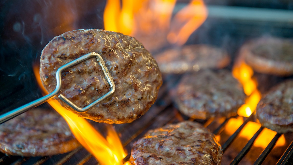 Tongs hold a burger over a flaming grill.