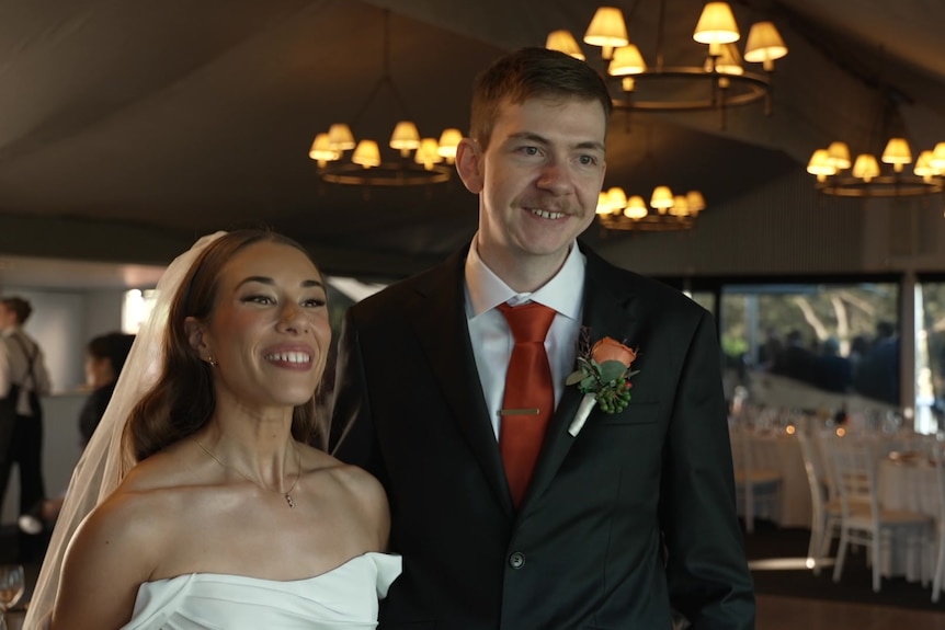 A woman in a wedding dress smiling next to a man in a suit with a wedding venue behind them.
